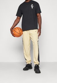 Schwarzes Baumwoll-T-Shirt mit weißem Grafikdesign, gelbe Sporthose mit schwarzen Seitenstreifen und schwarze Sneakers, hält einen orangefarbenen Basketball.