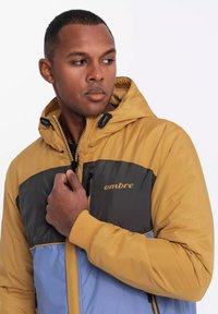 Färgat blockad pufferjacka i gul, grå och ljusblå. Har en dragkedja framtill, justerbar huva och elastiska ärmar. "ombre" logotypen visas.