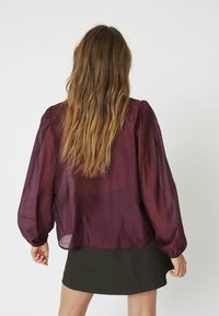 Blusa trasparente bordeaux con maniche a sbuffo, tessuto leggero e vestibilità ampia, abbinata a una gonna verde scuro. Vista posteriore.