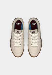 Ζευγάρι μπεζ Nike SB παπούτσια με λευκές κορδόνες, καφέ σόλες και μπορντώ εσωτερικούς πάτους τοποθετημένα το ένα δίπλα στο άλλο σε λευκό φόντο.