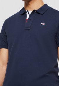 Mörkblå pikétröja i strukturvävt tyg, med en treknappsknäppning och en liten logotyp på bröstet i kontrasterande färger.