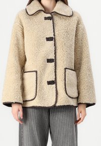 Beige shearling-takki ruskeilla reunuksilla, jossa on lovellinen kaulus, neljän napin suljenta ja kaksi etutaskua. Pehmeä, teksturoitu pinta.