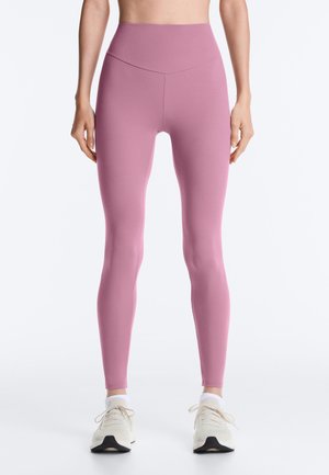 Žena v nohaviciach s vysokým pásom levandulovej farby a bielych teniskách stojí pred jednoduchým svetlým pozadím.