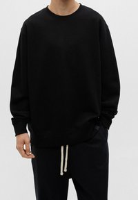 Svart sweatshirt med rund hals och lös passform, med ribbade muddar och fåll. Bärs med mörka byxor som har ljusa snören.