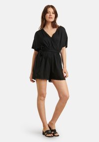 Black wrap romper features short sleeves, scalloped hem, and a tie belt. Soft fabric with a flowing design. Flat sandals complement the outfit.