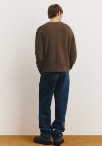 Barna bordázott, laza fazonú kereknyakú pulóver, sötétkék farmernadrággal és barna csizmával párosítva, hátulnézetben, egyszerű háttér előtt.