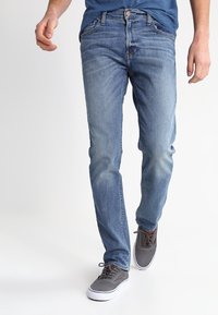 Jeans azul de corte slim con una textura desgastada, que cuentan con un cierre de botones y cinco bolsillos. Llevados con zapatillas gris, sobre un fondo claro.