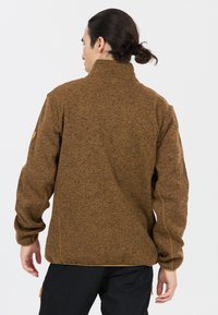 Giacca in pile marrone con colletto alto, design testurizzato e dettaglio con logo sulla parte superiore del braccio. Presenta una vestibilità rilassata e cuciture laterali.