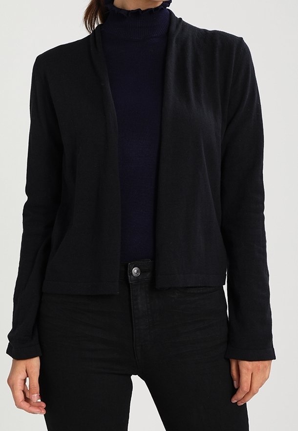 Černý cardigan s otevřeným předním dílem, texturovaným povrchem a dlouhými rukávy, kombinovaný s přiléhavým tmavým svetrem. Nosený přes černé džíny.