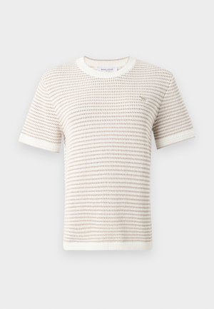 Femme portant un pull texturé blanc à manches courtes, un pantalon blanc large et des sandales noires et beiges, debout avec une main dans la poche.