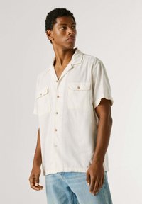 Jeune homme portant une chemise blanche ample à manches courtes avec deux poches poitrine, associée à un jean bleu clair, regardant sur le côté.