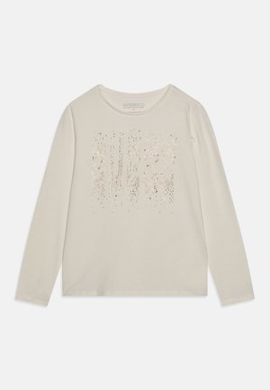 Chemise beige à manches longues avec col rond et "GUESS" écrit en petits clous métalliques sur la poitrine.
