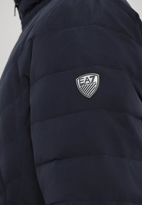 Jūrininkų pūkinė striukė, pasižyminti siuvinėtų raštų dizainu, minkšta tekstūra ir apsaugos logotipu kairiajame petyje, kontrastingos sidabrinės spalvos.