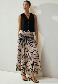 Sieviete, kas stāv pretī baltai sieni, ģērbusies melnā pogātā veste, beigu un melnā zebras raksta svārkos, kaklarotā un melnās strēmelēm ar augstiem papēžiem.