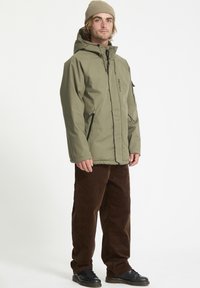 Veste isolante vert olive avec poches zippées, assortie avec un pantalon en velours côtelé marron et des chaussures noires à lacets. Un bonnet couvre les cheveux.