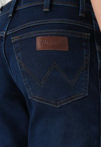 Jeans de mezclilla azul oscuro con un parche de cuero marrón que lleva la marca "Wrangler". Patrones de costura notables en los bolsillos traseros.