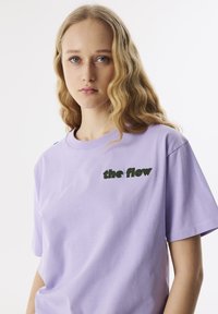 Lavendel katoenen t-shirt met korte mouwen en ronde hals. Heeft groene geborduurde tekst "the flow" op de linkerborst.