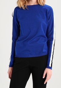 Pull en maille bleue à texture côtelée, avec des accents à rayures noires et blanches le long des manches, un col rond, et une coupe décontractée.