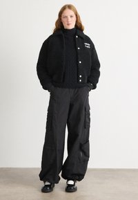 Veste courte noire et poilue avec boutons-pression et logo, associée à un pantalon cargo noir ample comportant des poches et des zips. Des chaussures noires complètent le look.