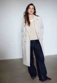 Lys grå uldfrakke med dobbeltknaplukning, parret med en beige sweater og mørkeblå vide jeans med kontrasterende syninger.