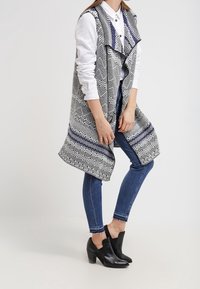 Gilet lungo in maglia bianco e nero con un motivo geometrico, caratterizzato da un design morbido e un collo a strati, abbinato a jeans blu e stivali alla caviglia.