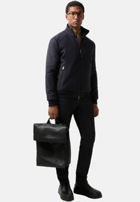 Marineblauer Reißverschluss-Jacke, schwarzer Leder-Rucksack, dunkle Jeans und schwarze Stiefel. Die Jacke verfügt über Reißverschlusstaschen und einen hohen Kragen.