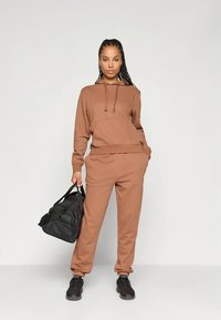 Camisola com capuz castanha com bolso canguru e cordão, combinada com calças de fato de treino a condizer. Mala desportiva preta e sapatos escuros completam o conjunto.