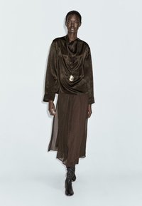 Braun drapiertes Oberteil mit einem Carmenausschnitt, kombiniert mit einem langen, transparenten braunen Rock und knielangen schwarzen Stiefeln, die minimale Details sowie eine Anhänger-Halskette aufweisen.