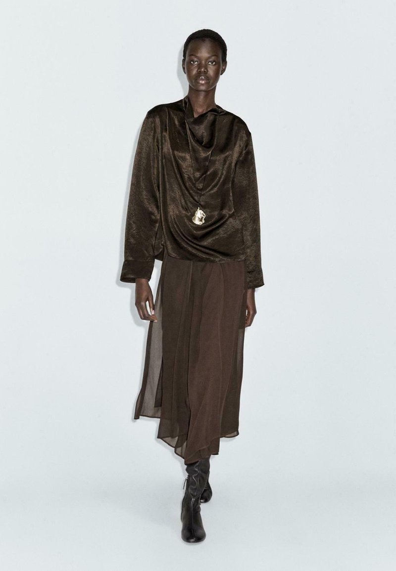 Braun drapiertes Oberteil mit einem Carmenausschnitt, kombiniert mit einem langen, transparenten braunen Rock und knielangen schwarzen Stiefeln, die minimale Details sowie eine Anhänger-Halskette aufweisen.