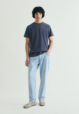 Junger Mann mit lockigem dunklem Haar trägt ein dunkelblaues T-Shirt, hellblaue Jeans und graue Sneakers, steht mit einer Hand in der Tasche.