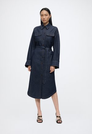 Kvinde iført en mørk denim midi-knapkjole med lange ærmer, brystlommer, bindebånd i taljen og sorte højhælede sandaler med stropper.