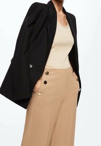 Zwarte blazer met gevoerde schouders, gecombineerd met een beige tanktop en hoge beige broeken met grote goudkleurige knopen.