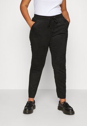 Person wearing black drawstring pants and black loafers standing against a plain light background, hands in pockets.