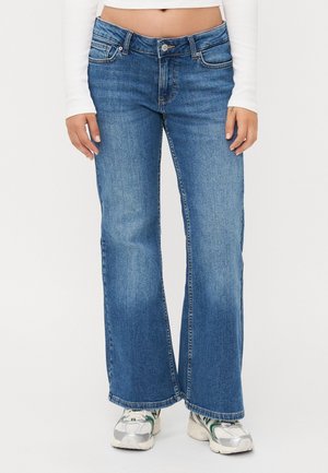 Kobieta ubrana w niebieskie jeansy bootcut, biały długi crop top i białe trampki z zielonymi akcentami, stojąca na jednolitym tle.