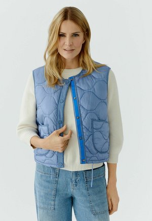 Femme portant un gilet matelassé bleu clair par-dessus un pull blanc et un jean bleu, debout devant un fond clair uni.