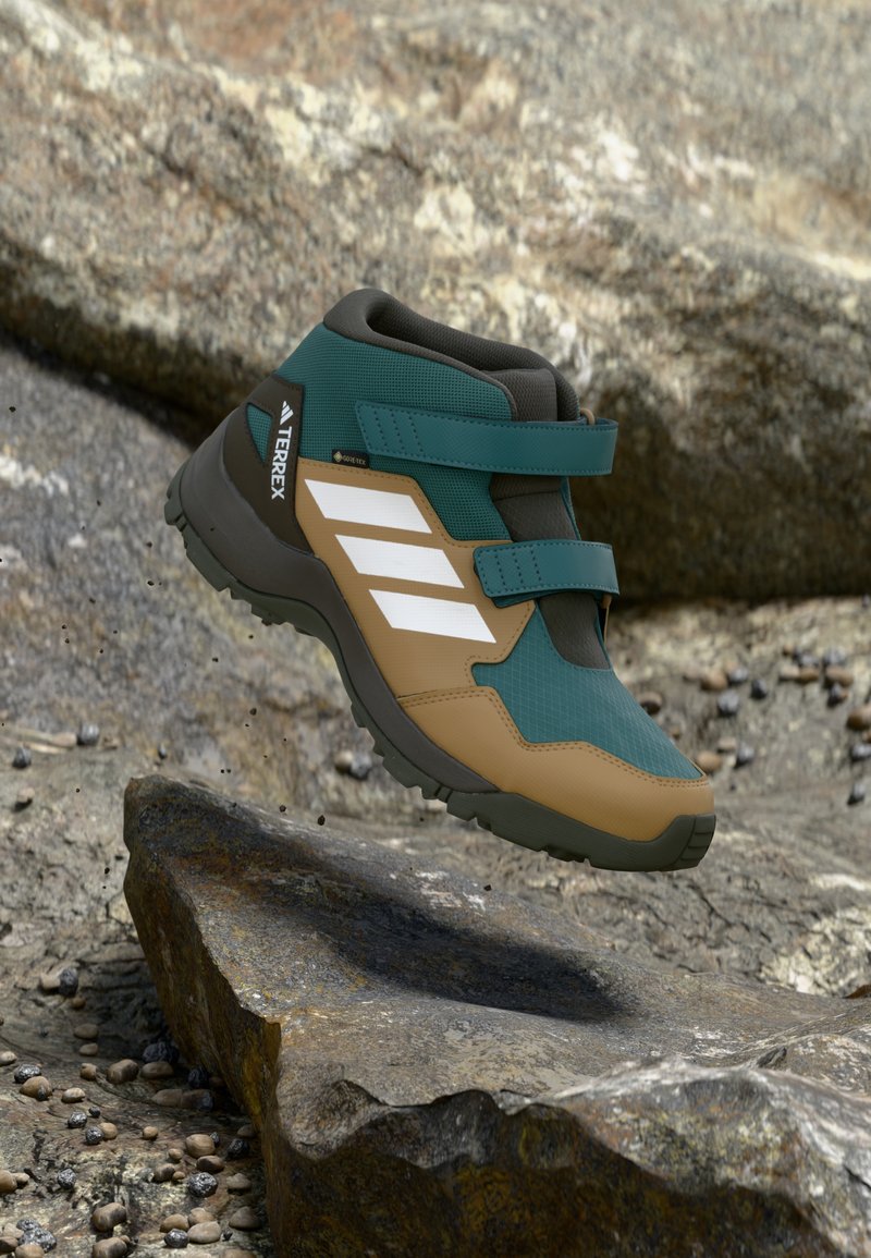 High-top green and tan hiking shoe with Velcro straps, textured synthetic upper, rubber sole, and three white stripes on the side.