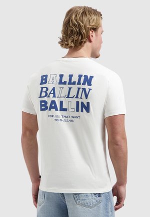 Man met blond haar, met de rug naar de camera, draagt een wit T-shirt met blauwe tekst "BALLIN BALLIN BALLIN" en kleinere tekst eronder, lichtblauwe spijkerbroek.