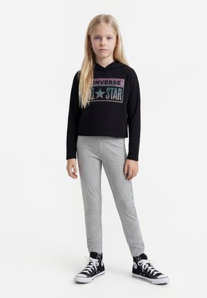 Szőke lány áll, fekete Converse All Star kapucnis pulóvert, szürke szabadidőnadrágot és fekete magas szárú Converse tornacipőt viselve, fehér háttér előtt.