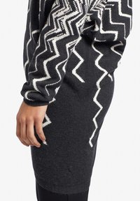 Czarna szara sukienka z długim rękawem z białymi wzorami w zygzak na rękawach i bokach, noszona przez osobę z ręką swobodnie opadającą wzdłuż ciała.