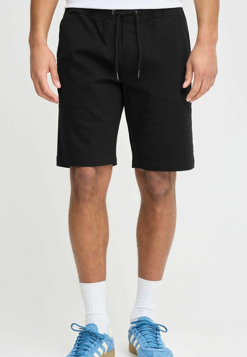 Svarte bomullsshorts med elastisk midjebånd og justerbar snor, med en avslappet passform og rullet kant, brukt med blå sneakers.
