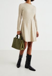Abito lavorato a maglia beige con texture a coste, vita arricciata, abbinato a una borsa a tracolla in pelle verde e stivaletti alla caviglia in pelle nera.