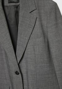 Blazer gris pour homme à un bouton, avec revers crantés et petite poche poitrine, révélant la doublure intérieure noire et l'étiquette de la marque.
