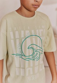 Jasnozielona bawełniana koszulka z grafiką przedstawiającą falę oraz napisem "CATCH SOME WAVES" w kontrastowych białych i turkusowych kolorach.