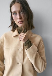 Lysebeige ruskindsjakke med krave, der har to runde knapper og en glat tekstur. Båret over en mørkegrøn rullekrave.