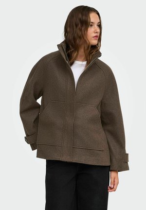 Jeune personne portant une veste zippée marron avec un col montant et des poches, associée à un pantalon noir, regardant sur le côté sur un fond uni.