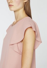 Donna che indossa un top senza maniche di un delicato rosa chiaro con maniche a strati e a leggerezza, visibile dal collo fino al braccio superiore su uno sfondo semplice.