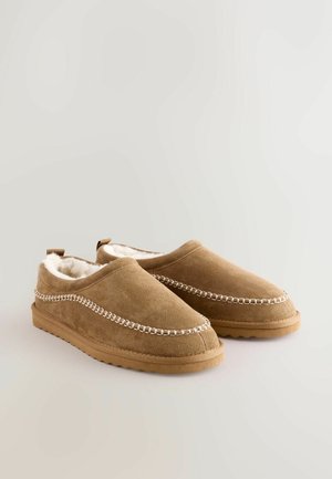 Zapatos de ante color marrón claro sin cordones con suave forro de peluche sintético, costuras decorativas en blanco a lo largo de los lados y suelas de goma antideslizantes.