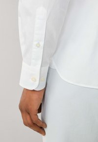 Camisa blanca de manga larga con puños abotonados, que presenta una textura suave, un diseño de puño puntiagudo y costuras de acento en azul claro cerca de un botón.