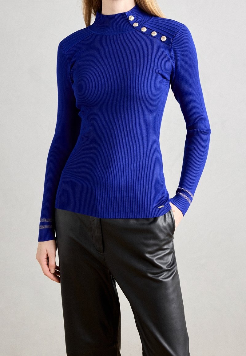 Femme portant un pull côtelé bleu roi ajusté avec des boutons métalliques sur l'épaule et un pantalon en cuir noir, main dans la poche.