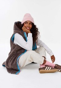 Fleece vesta v tmavě hnědé barvě s tyrkysovou podšívkou, spárovaná s bílým dlouhým rukávem a béžovými kalhotami. Světle růžová čepice a ponožky. Hnědé boty.